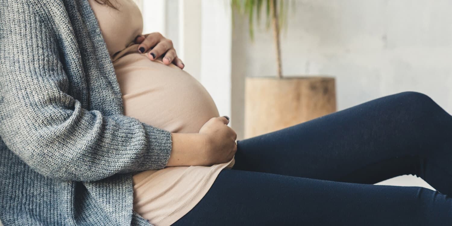 Pregnant woman sits on a blanket on the floor and clasps her baby bump with both hands.