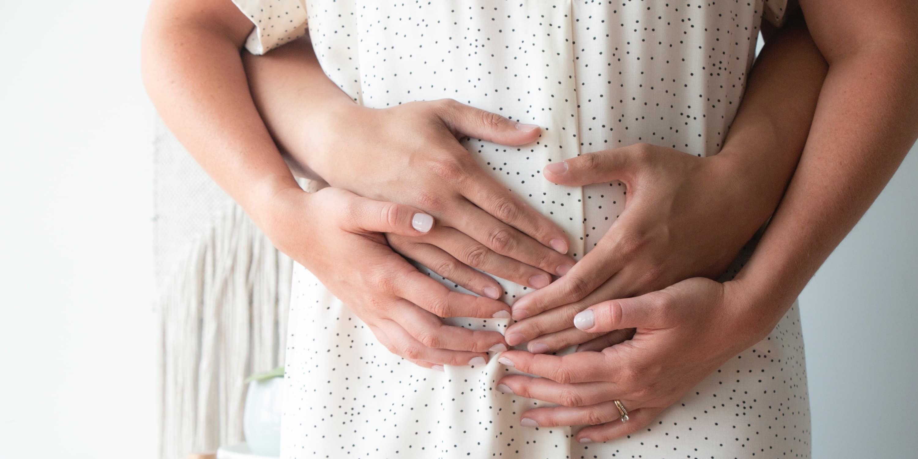 Een man omarmt een vrouw van achteren op haar buik en de vrouw legt haar handen op die van hem.