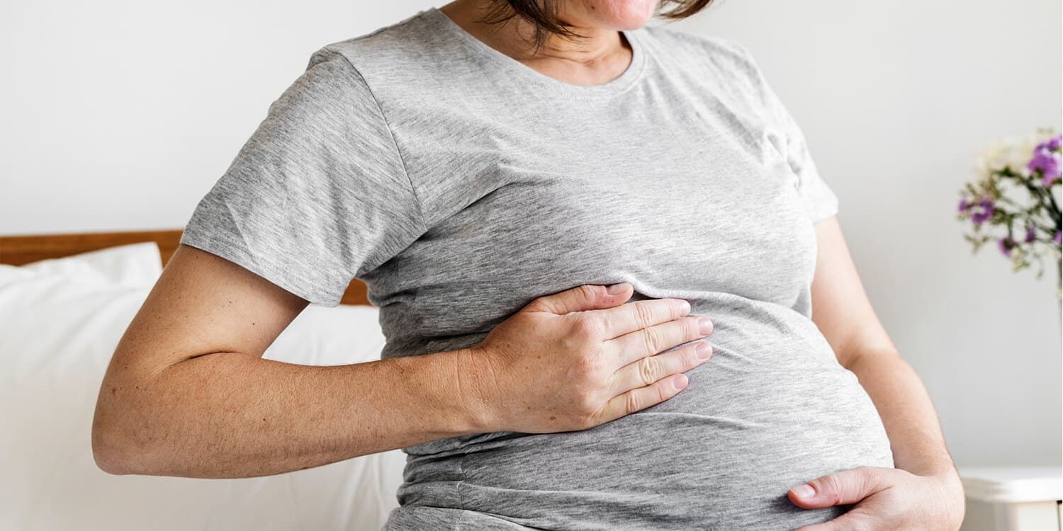 Gravid kvinne sitter i sengen, kjenner på magen og smiler.