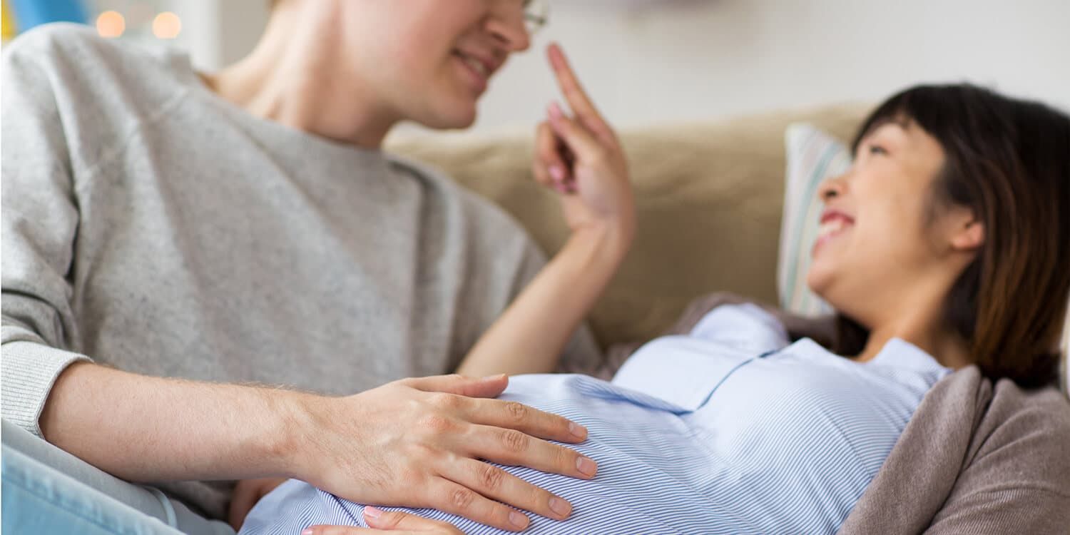 casal grávido rindo aninhado no sofá, homem coloca a mão na barriga de grávida da mulher