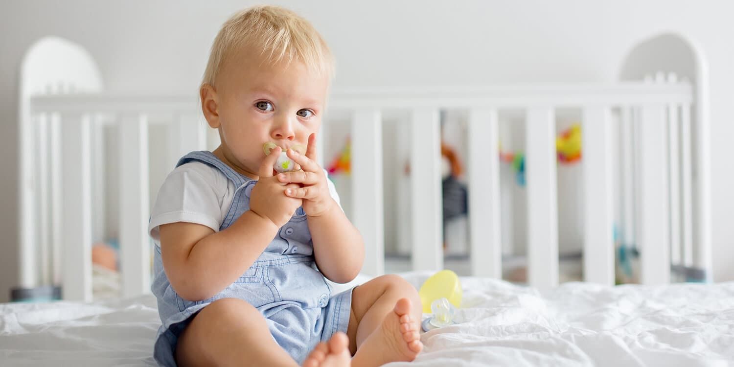 Baby mit Schnuller sitzt in Bett und hält den Schnuller mit beiden Händen fest