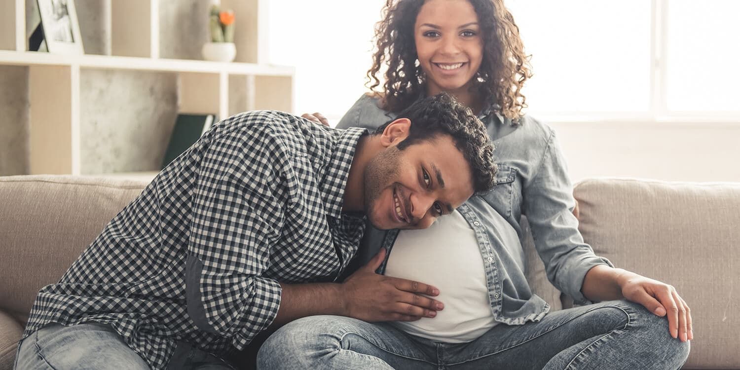 Een man en een zwangere vrouw zitten op de bank, waarbij de man zijn oor op de buik van de vrouw legt.