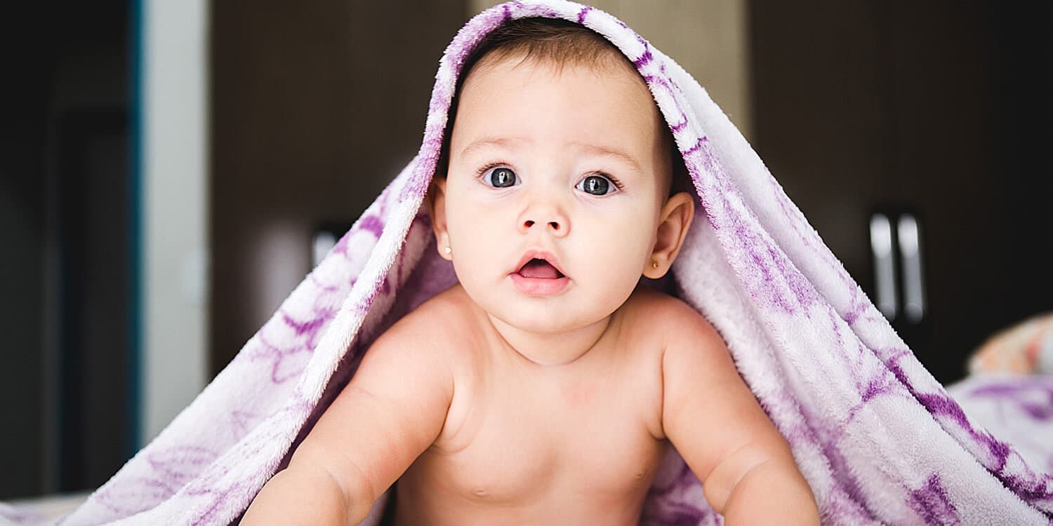 Baby liegt auf dem Bauch, hat ein Handtuch über dem Kopf und blickt in die Kamera