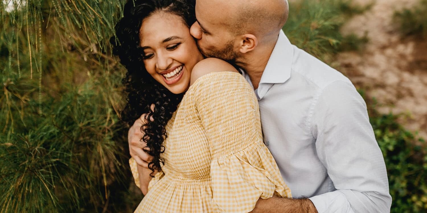 Man has his arms around his pregnant partner and is kissing her on the cheek 