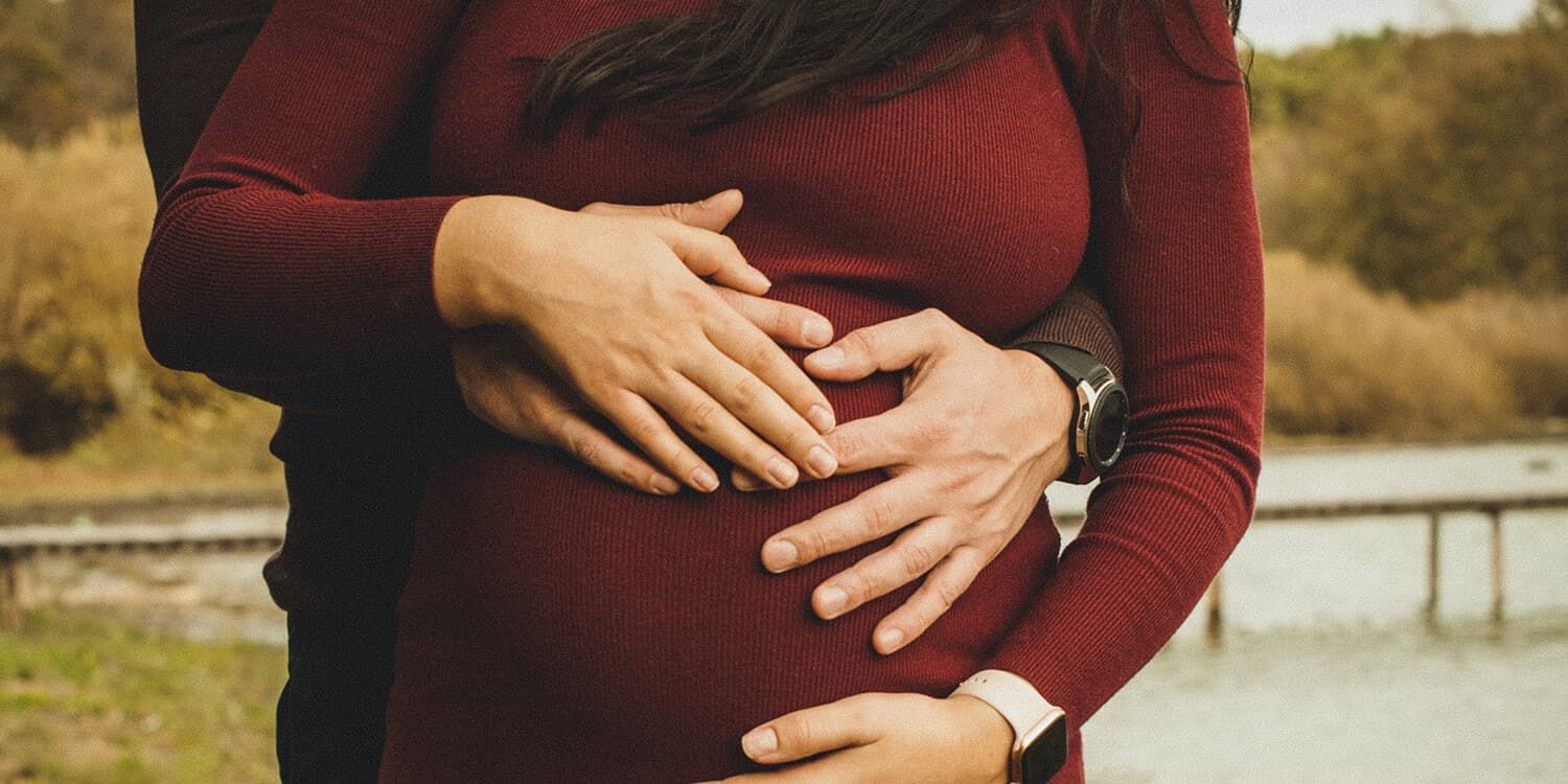 Casal grávido ao ar livre; ambos abraçam a barriga da mulher 