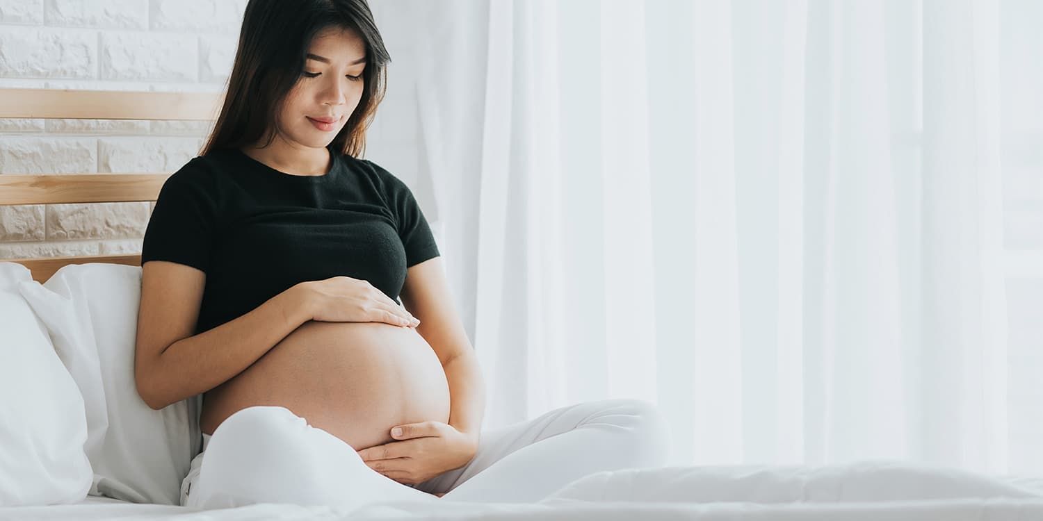 Gravid kvinne sitter på sengen og omfavner babymagen forsiktig.