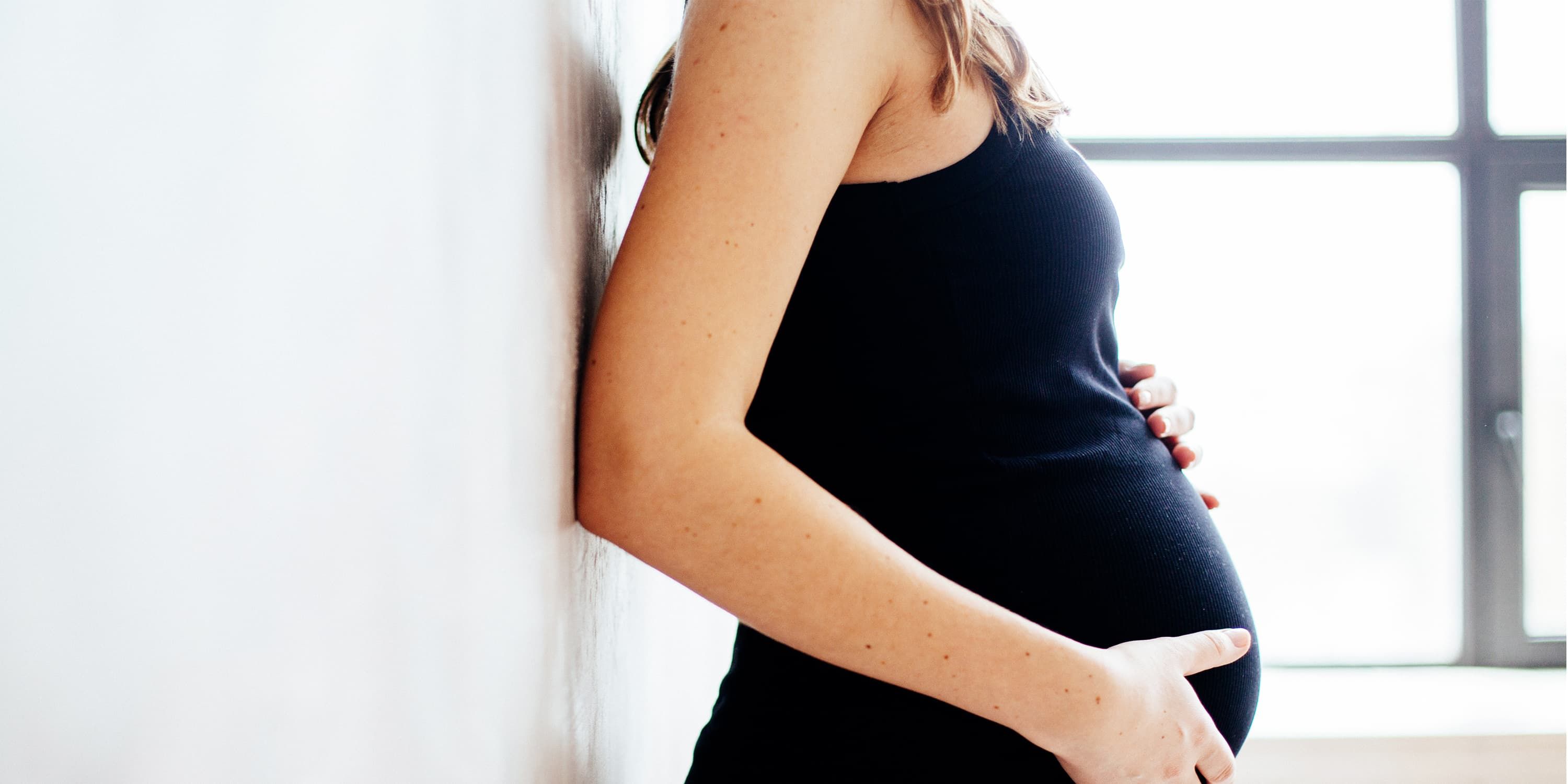 Seitenansicht, schwangere Frau lehnt an der Wand und legt die Hände auf ihren Bauch